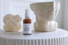 Load image into Gallery viewer, Mist by Simple Rituals a frosted white bottle with a spray cap containing pink liquid with a cream bust of a woman cut off at the eyes and a cream Celtic cross candle behind 
