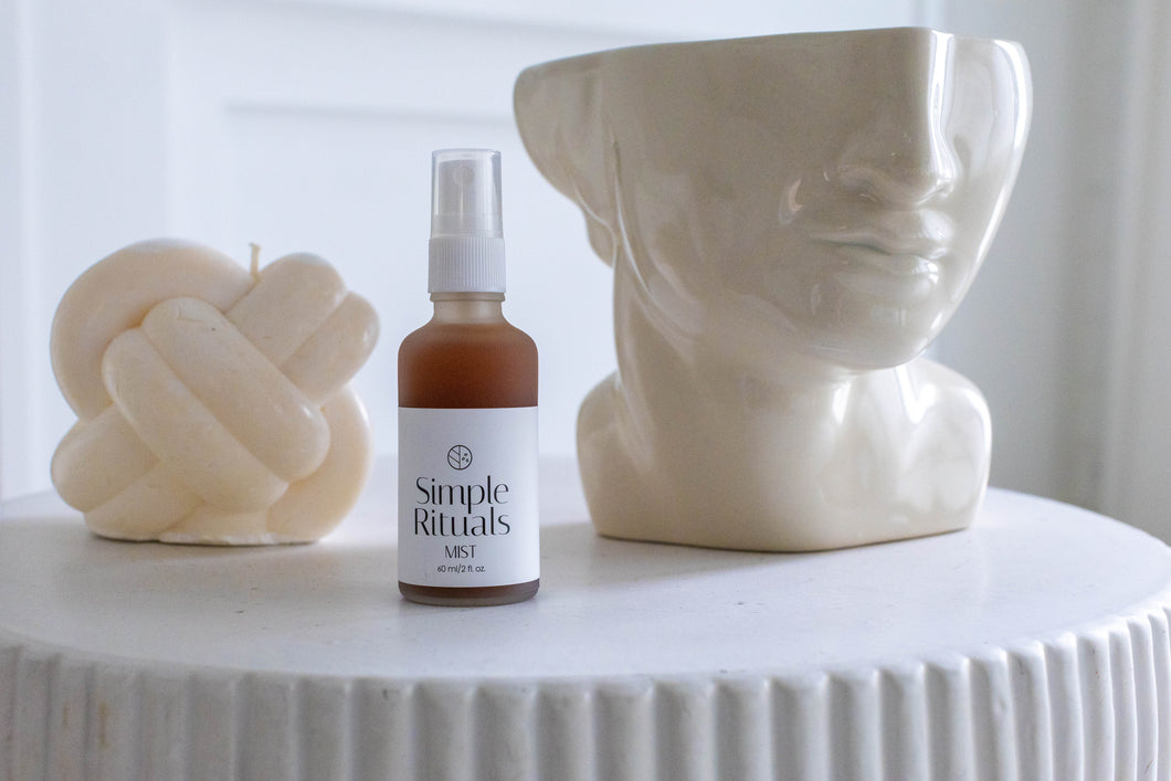 Mist by Simple Rituals a frosted white bottle with a spray cap containing pink liquid with a cream bust of a woman cut off at the eyes and a cream Celtic cross candle behind 