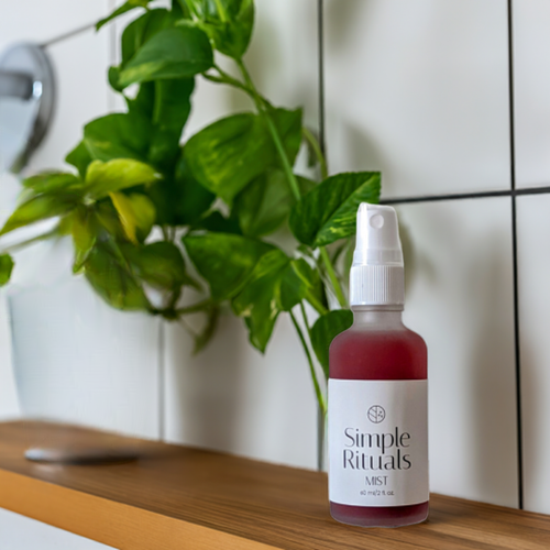 Spritz bottle of Mist. A pink liquid in a frosted glass bottle on a wood shelf with white tiled walls behind and a plant hanging down.